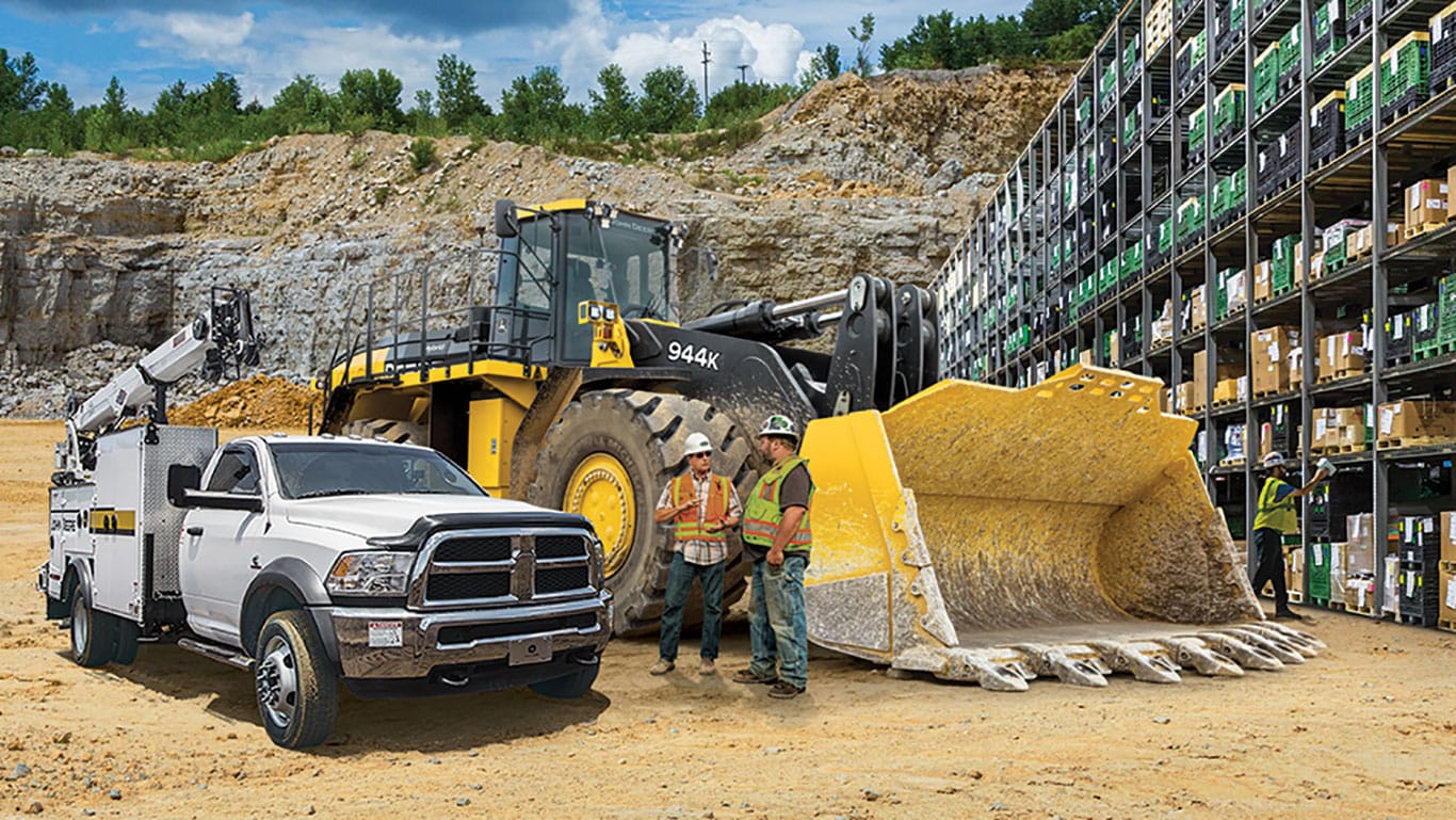 two construction workers standing in front of a John Deere 944K wheel loader with a shelf of John Deere construction equipment parts behind them.